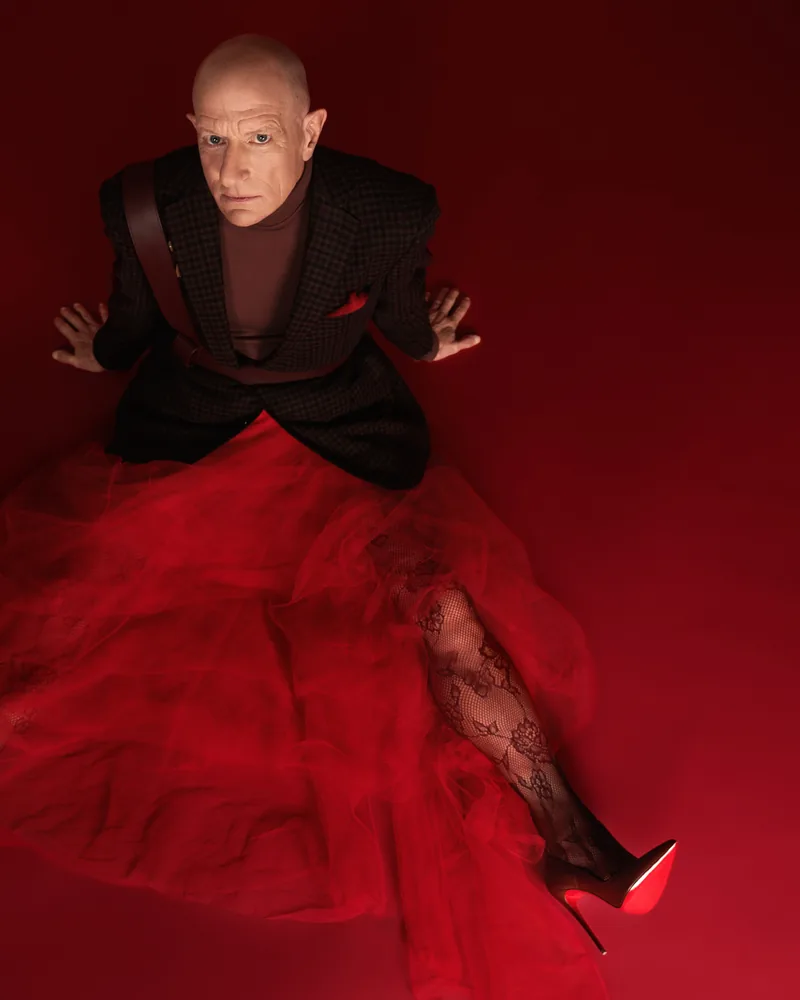 Fashion portrait of a seated figure in a dark jacket and a voluminous red tulle skirt with a red stiletto, on a deep red ground